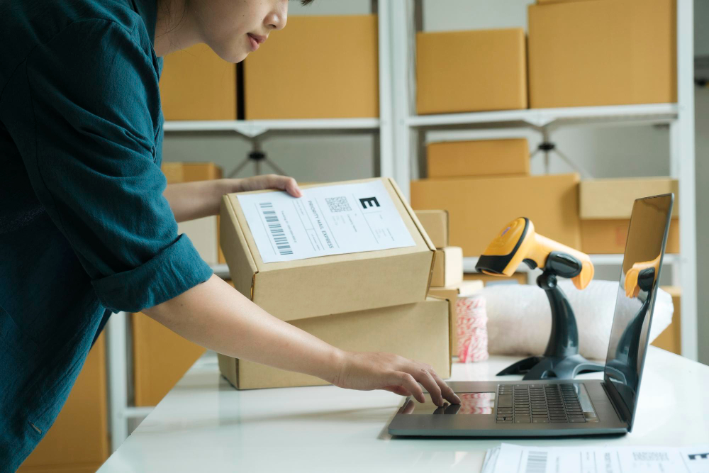 A person packaging a box for shipping, scanning barcodes and entering data on a laptop, with cardboard boxes stacked in the background