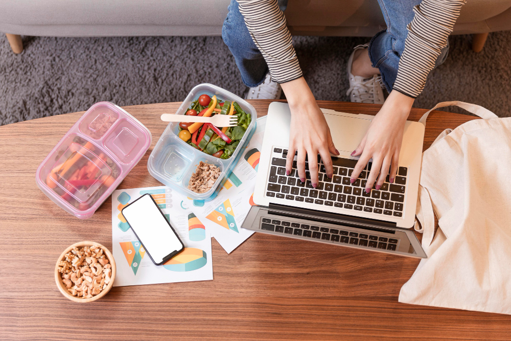 A person typing on a laptop at a wooden table surrounded by healthy meal prep containers, a smartphone, printed product mockups, and a bowl of cashews