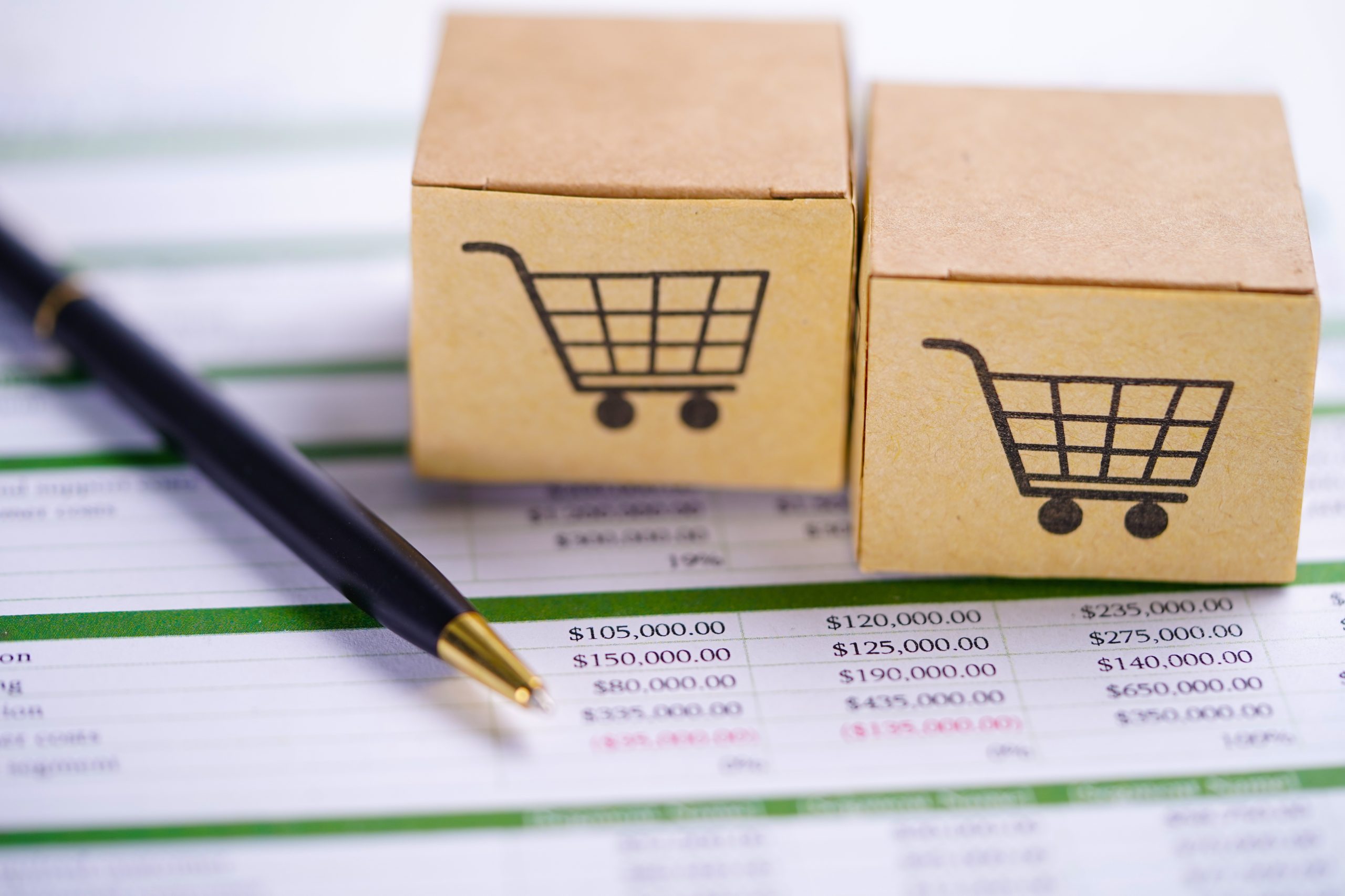 personalized pricing - Wholesale Suite Two small cardboard boxes with shopping cart icons placed on top of a financial spreadsheet, alongside a black and gold pen