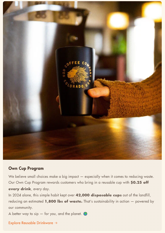Hand holding a black reusable OZO Coffee Company cup inside a cozy café environment as part of a sustainability initiative.