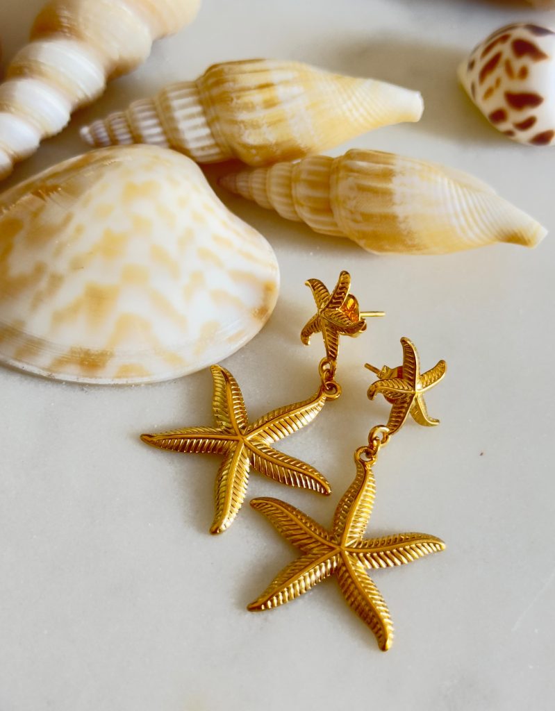 Gold starfish earrings displayed beside assorted seashells on a light surface