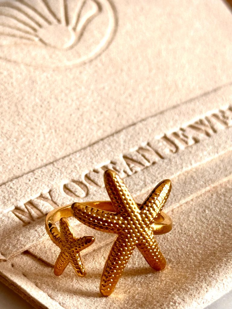 Gold double starfish ring displayed on a soft beige jewelry pouch with embossed "MY OCEAN JEWELLERY" branding in the background.