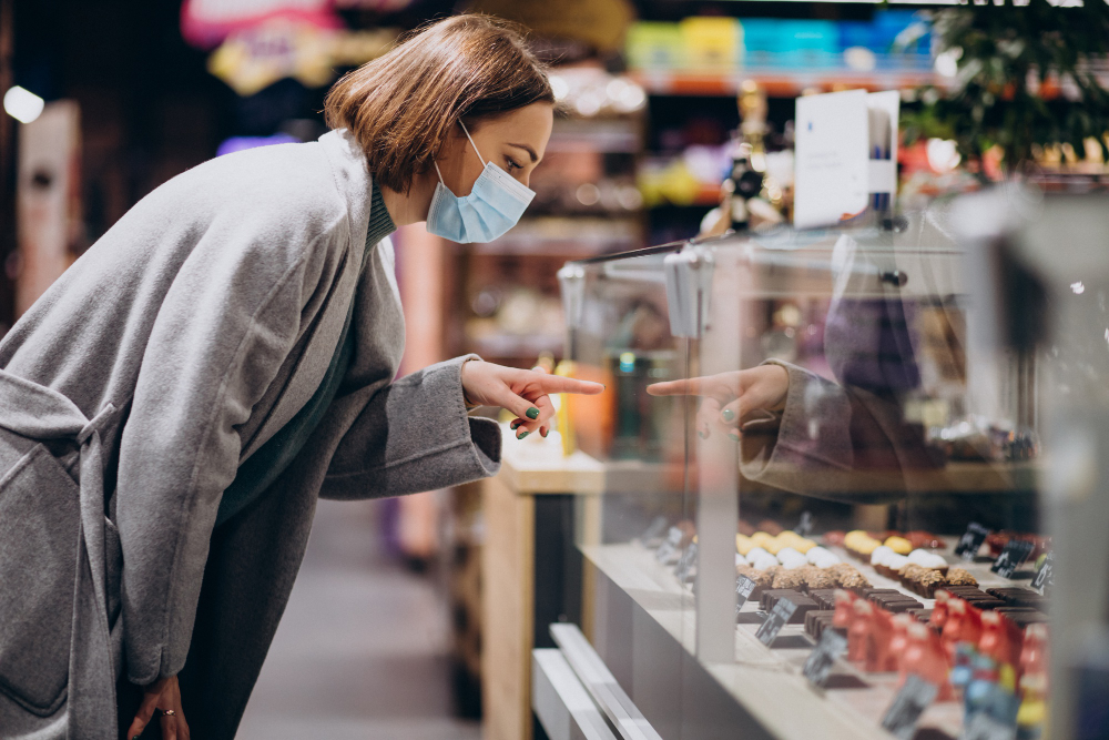 Woman wearing a face mask and winter coat points at assorted chocolates behind a glass counter in a retail store