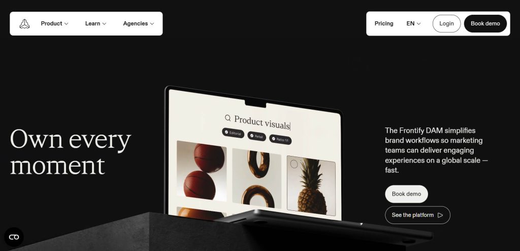 Dark-themed webpage showing a tilted laptop screen displaying a product visuals grid, flanked by bold 'Own every moment' headline and demo call-to-action.